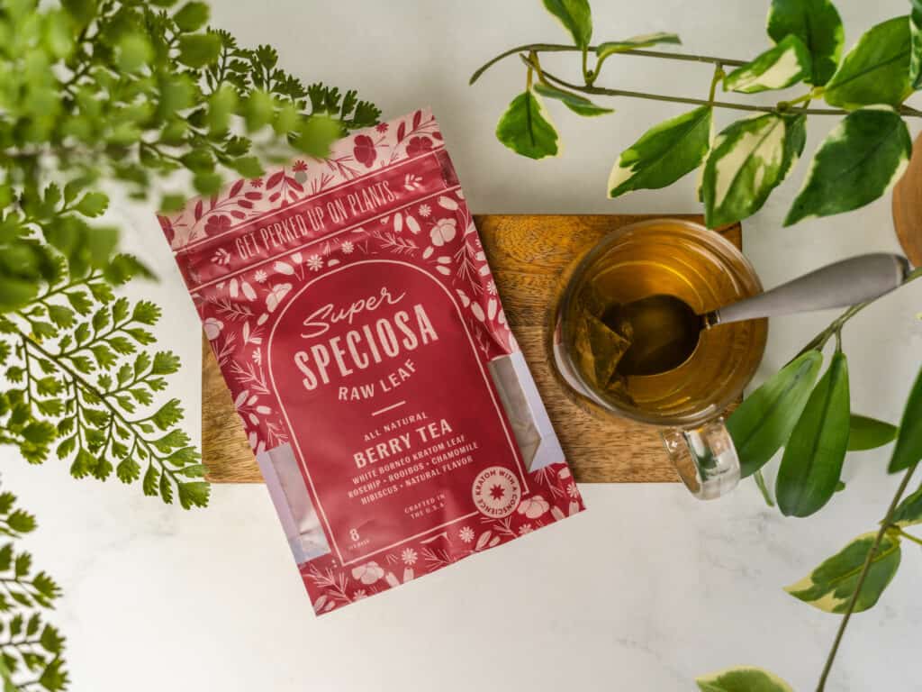 Super Speciosa Berry Kratom Tea with cup and tea bag and tropical leaves. Herbal tea blend for energy.