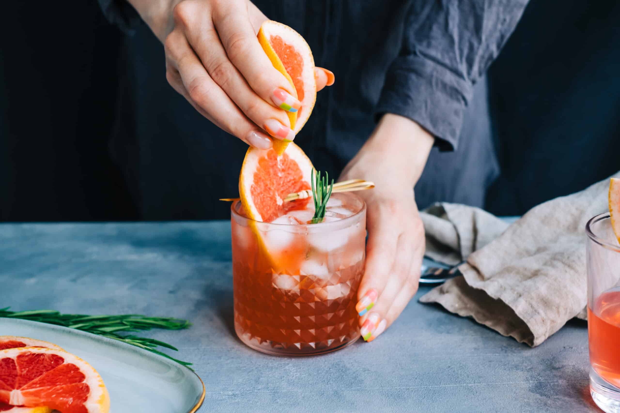 Female bartender hand squeezes juice from fresh grapefruit in cocktail lemonade with ice and rosemary. Grapefruit is also used a kratom potentiator.